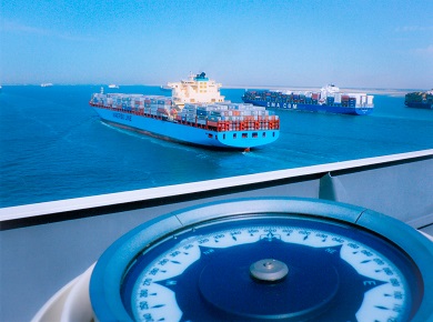 Large compass binnacle in foreground with a view of a freighter boat traversing the ocean in the background