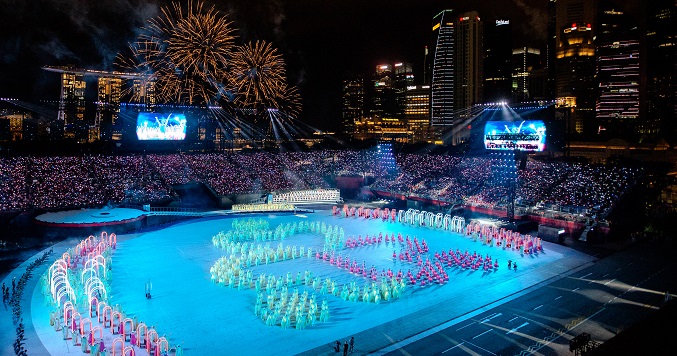 Una coreografía en un campo de juegos rodeado de público con fuegos artificiales de fondo