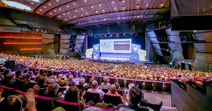 People seated in a large auditorium looking at the stage where a presentation is taking place during a conference.