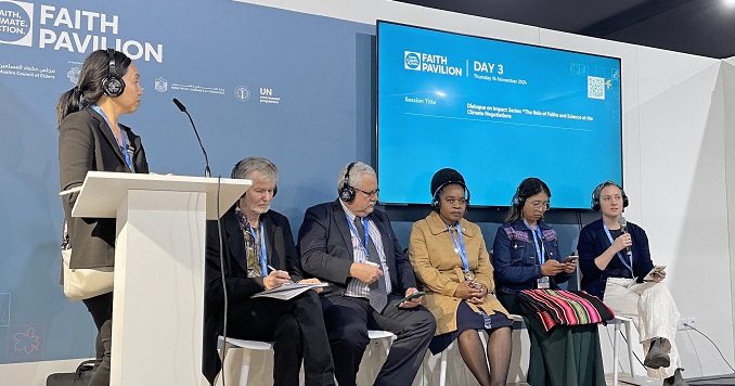 A six-person panel sits on stage, with one individual standing at a central podium, while the panelist seated at the far right speaks into a microphone.