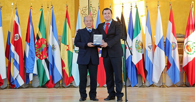 Two men standing in front of flags holding a medal.