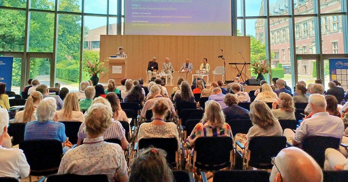 Numerosas personas sentadas en una sala de conferencias frente al escenario en una sala con grandes ventanales.