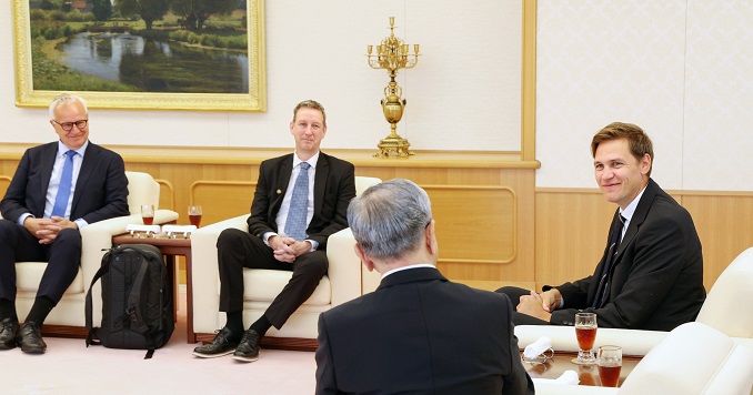 Four men in smart suits engage in conversation in a large room with white armchairs. 