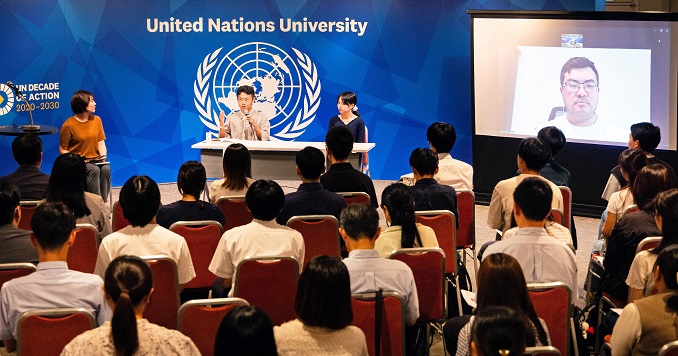 A panel of three speakers on stage and one on screen engaged in discussion before a small audience. 