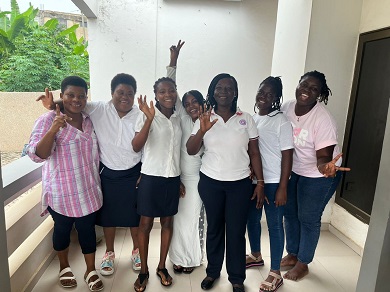 A group of young women standing in front of a building, smiling and waving.
