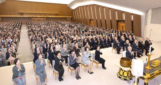 Many people sitting in rows in a large hall praying.