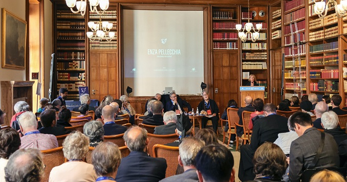 A woman sitting at the front of a room lined with bookshelves with an audience looking on.