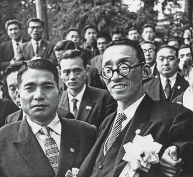 Close-cropped image of Ikeda and Toda facing the camera at the front of a group of men