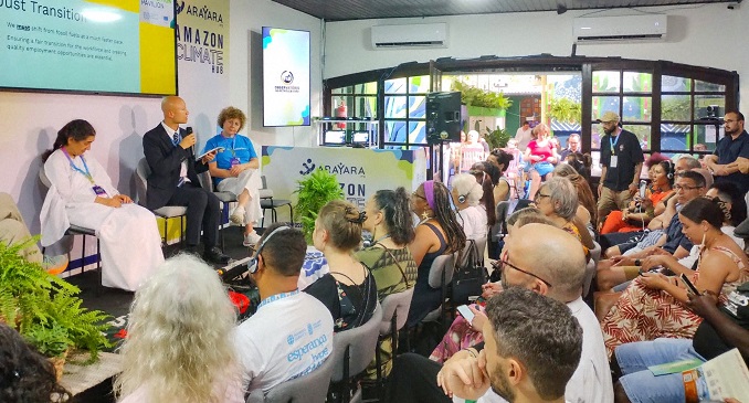 Three people sit on a small stage and engage in conversation with an audience in a conference room. 