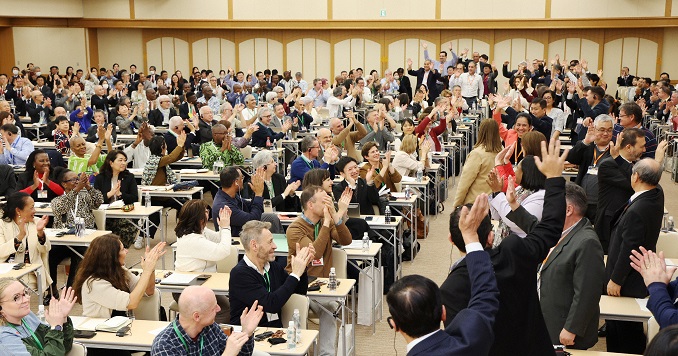 Many people in a large hall sitting at desks turning to the back of the room and waving to a small group entering the room.