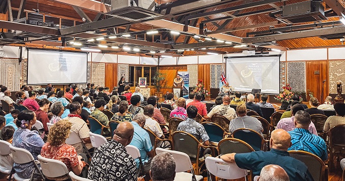 Audience members seated in a large conference hall listen to a speaker on stage.