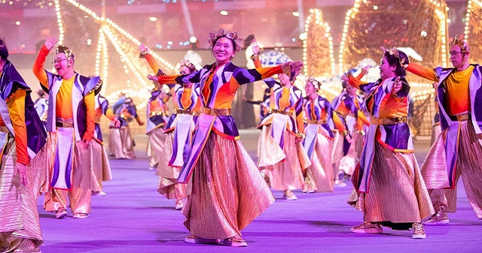 Performers in colorful costumes dance on the stadium stage, with large, illuminated boat-shaped structures behind them.