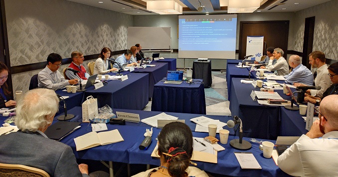 People seated around tables on three sides of a small conference room, with a presentation screen at the front.
